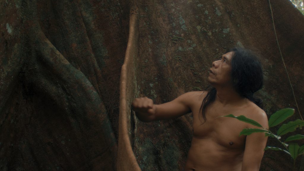 A still from episode 8 of Leviathan, shows an South American indigenous man, pounding a tree with his fist, looking up at it.