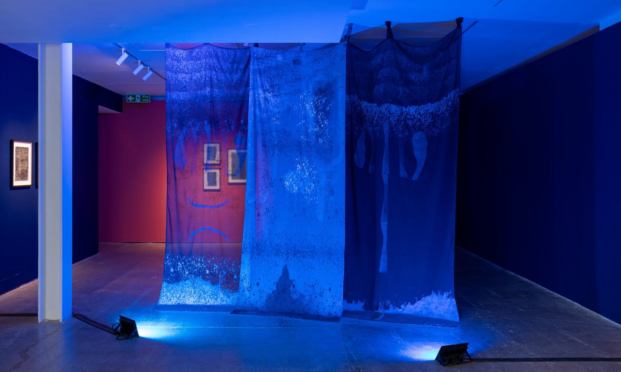 Photograph showing Jesica El Mal's work installed at Castlefield Gallery. Three transulsent blue banners, lit by two blue lights, hang floor to celing infront of a red wall.