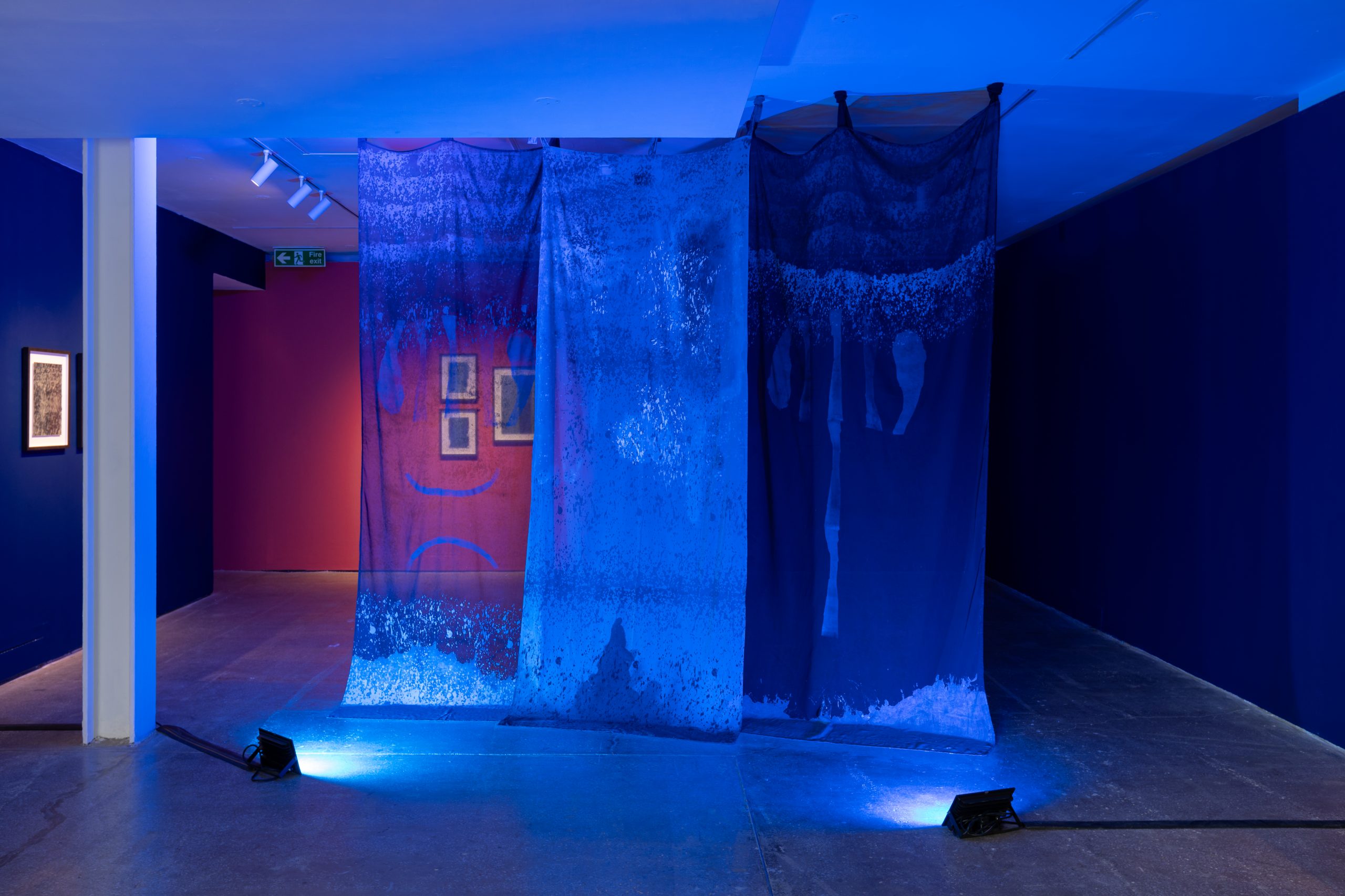 Photograph showing Jesica El Mal's work installed at Castlefield Gallery. Three transulsent blue banners, lit by two blue lights, hang floor to celing infront of a red wall.
