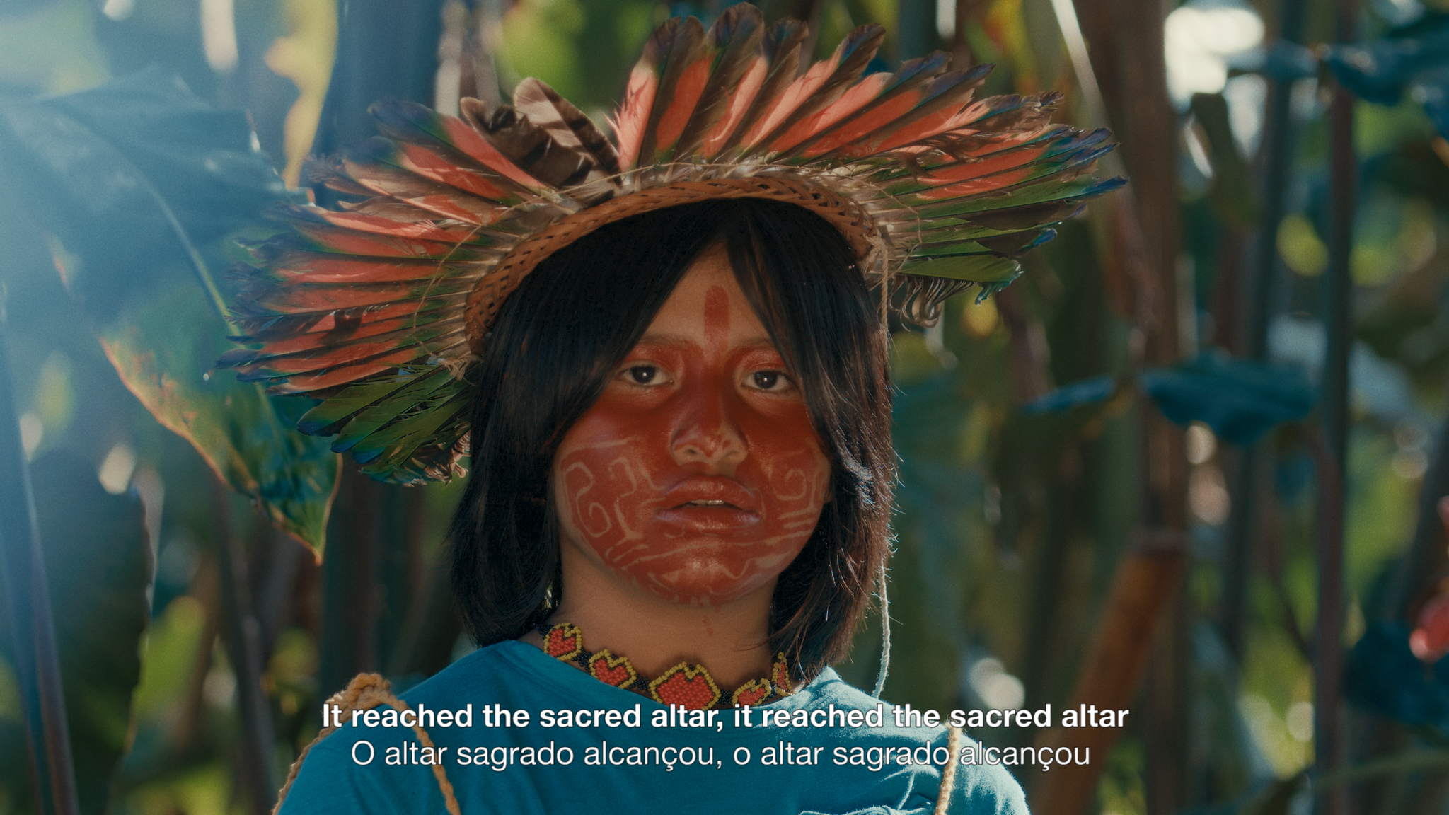A still image from Leviathan Episode 8 showing a young indigenous person in traditional dress and face paint looks into the camera. A caption included on the image reads: 'It reached the sacred alter, It reached the sacred alter".