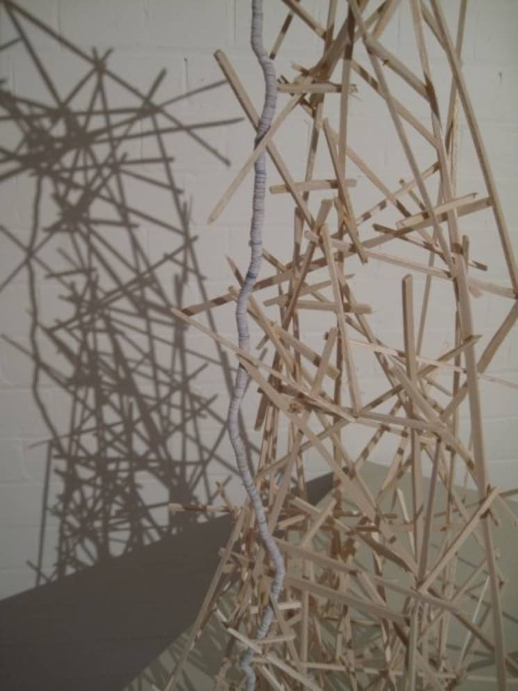 A sculptural work made of many small sticks of pale wood connected precariously. The work casts a dramatic shadow.